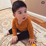 Imran is registered to the contest to win money with this photo: animal_print, black_pants, boy, carpet, child, curious, door, face, home, indoors, looking_up, orange_shirt, portrait, rug, sitting, toddler, toy, wall, white_sock, wood_floor
