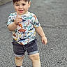 child, toddler, boy, smiling, thumbs_up, portrait, outdoor, standing, fashion, dinosaur_shirt, shorts, sneakers, hair, face, road, pavement, park, daylight, happy, photography