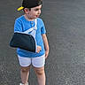 child, boy, outdoor, portrait, standing, blue_shirt, baseball_cap, backward_cap, arm_sling, injured_arm, shorts, sandals, pavement, street, road, greenery, daylight, profile, casual_clothing, person