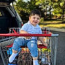 child, toddler, boy, smiling, sitting, shopping_cart, grocery_cart, outdoors, parked_car, trees, street, sunlight, blue_shirt, blue_jeans, white_socks, vehicle, portrait, happy, shopping, daytime