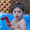 backyard, blue_water, child, close_up, dark_hair, face, outdoor, photograph, playtime, pool, portrait, red_toy, serious_expression, summer, swimming, toddler, toy_gun, water, water_blaster, wet