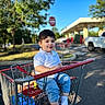 child, boy, smiling, shopping_cart, outdoor, parking_lot, supermarket, trees, blue_sky, sunlight, casual_clothing, white_shirt, jeans, socks, portrait, face, vehicle, stop_sign, cart, playful
