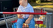 Imran is registered to the contest to win money with this photo: child, toddler, boy, shopping_cart, outdoor, parked_car, grocery_shopping, sitting, portrait, profile, short_hair, white_shirt, blue_jeans, white_socks, sunlight, trees, grass, vehicle, retail, family_outing