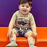 child, toddler, baby, smiling, happy, portrait, sitting, orange_couch, purple_background, dinosaur_shirt, shorts, socks, shoes, toy, indoor, colorful, face, person, casual_clothing, photography