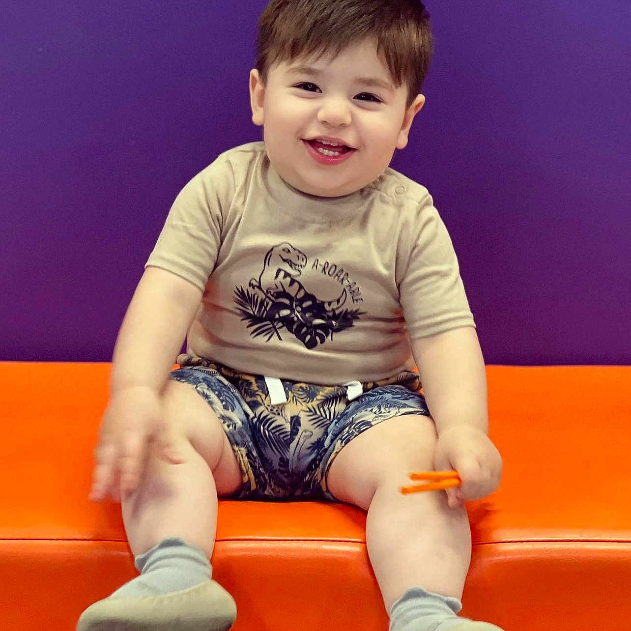 Imran is registered to the contest to win money with this photo: child, toddler, baby, smiling, happy, portrait, sitting, orange_couch, purple_background, dinosaur_shirt, shorts, socks, shoes, toy, indoor, colorful, face, person, casual_clothing, photography