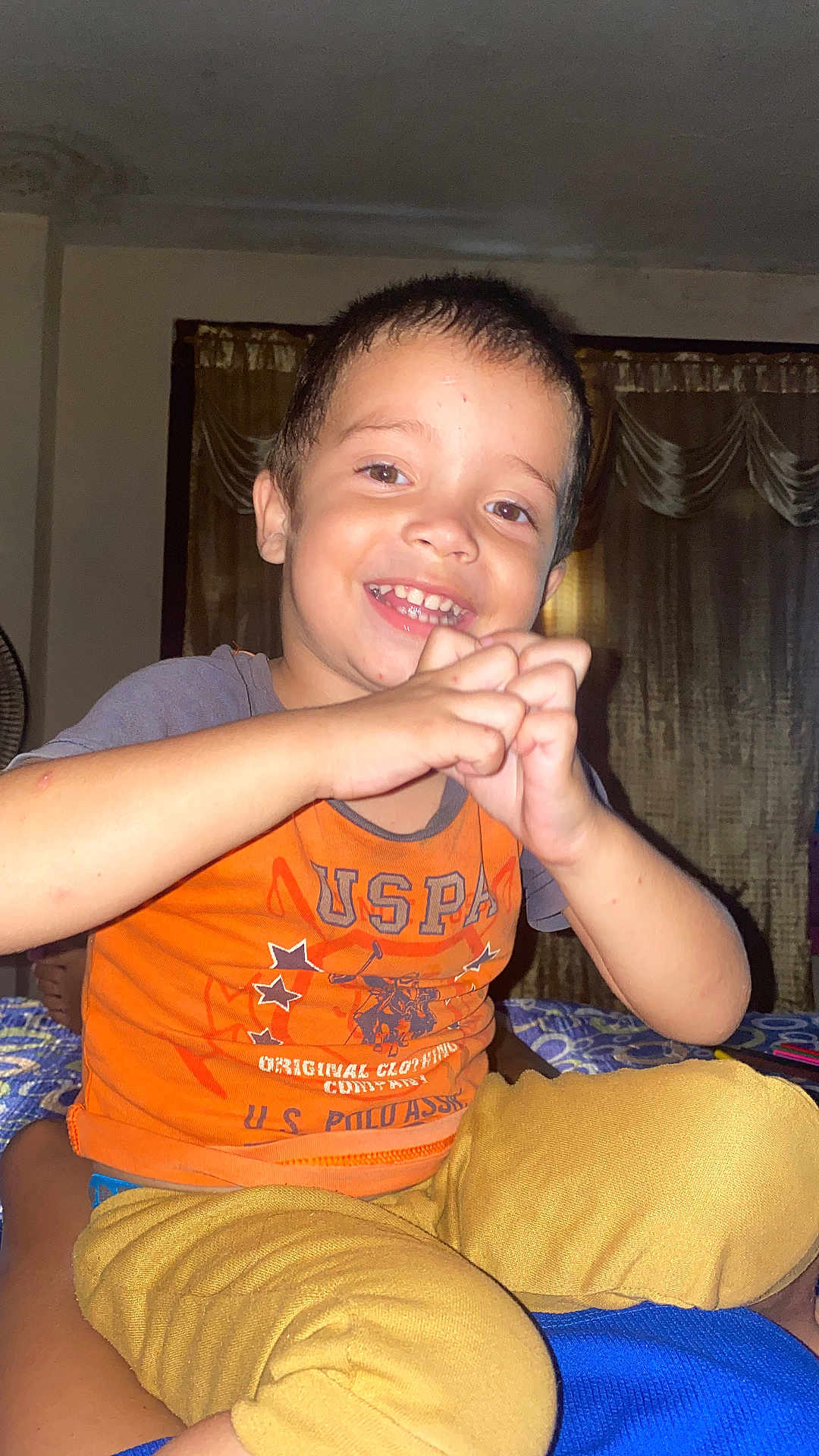 Lukas joined the competition — help win amazing prizes! child, boy, smile, hands, orange_shirt, mustard_pants, indoor, curtains, fan, happy, person, face, sitting, casual_clothing, playful, young, portrait, home, expression, cute