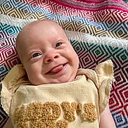 Scottie is registered to the contest to win money with this photo: baby, infant, smile, tongue_out, face, blanket, patterned_blanket, colorful, cute, happy, clothing, yellow_outfit, closeup, portrait, child, young, skin, head, eyes, expression