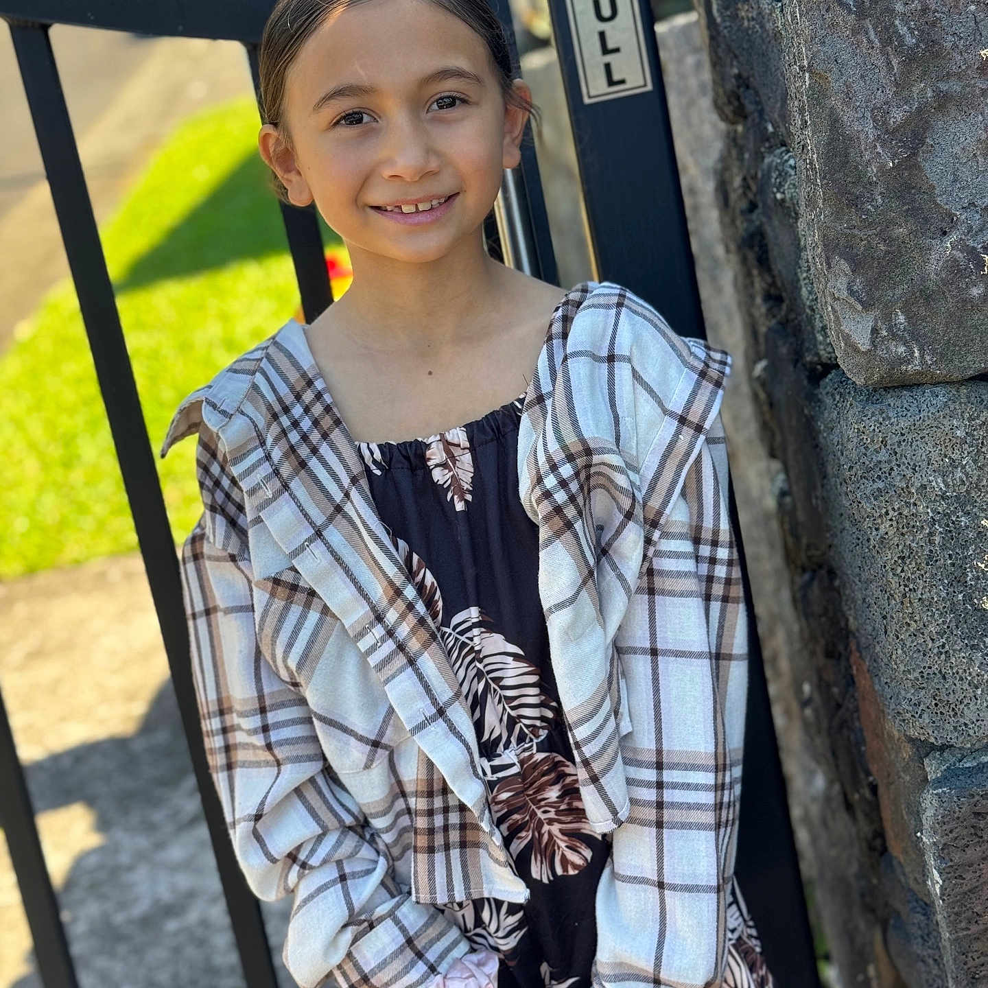 Kyleiha Marie is registered to the contest to win money with this photo: casual, child, clothing, daylight, dress, face, gate, girl, hair, happy, nature, outdoor, person, plaid_shirt, portrait, smiling, standing, stone_wall, sunlight, young