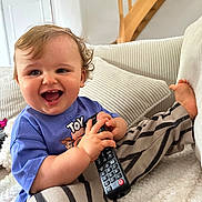 Sacha participe au concours pour gagner de l'argent avec cette photo : baby, child, remote_control, smiling, curly_hair, purple_shirt, striped_pants, couch, cushion, indoor, wooden_stairs, white_wall, happy, sitting, toy_story, person, foot, hand, cozy, home