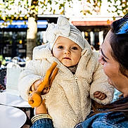 Lya participe au concours pour gagner de l'argent avec cette photo : baby, child, woman, outdoor, cafe, toy, fluffy_jacket, headband, smile, holding, person, face, daylight, table, cup, chair, people, trees, background, fashion