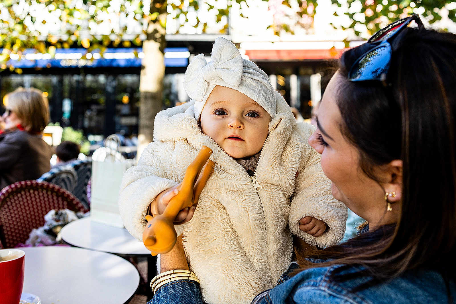 Lya participe au concours pour gagner de l'argent avec cette photo : baby, child, woman, outdoor, cafe, toy, fluffy_jacket, headband, smile, holding, person, face, daylight, table, cup, chair, people, trees, background, fashion