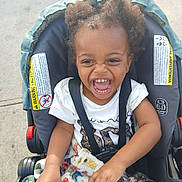 Quilan joined the competition — help win amazing prizes! toddler, child, stroller, laughing, curly_hair, blanket, white_tshirt, outdoor, sunny, sidewalk, happy, baby, seatbelt, cute, joyful, person, casual_clothing, smile, playful, portrait