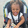 toddler, child, stroller, laughing, curly_hair, blanket, white_tshirt, outdoor, sunny, sidewalk, happy, baby, seatbelt, cute, joyful, person, casual_clothing, smile, playful, portrait