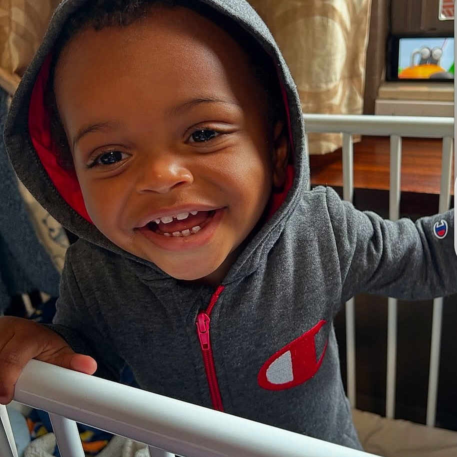 Quilan is registered to the contest to win money with this photo: toddler, child, hoodie, smiling, crib, indoor, curtain, happy, baby, clothing, person, furniture, toy, railing, face, portrait, young_child, home, cute, casual