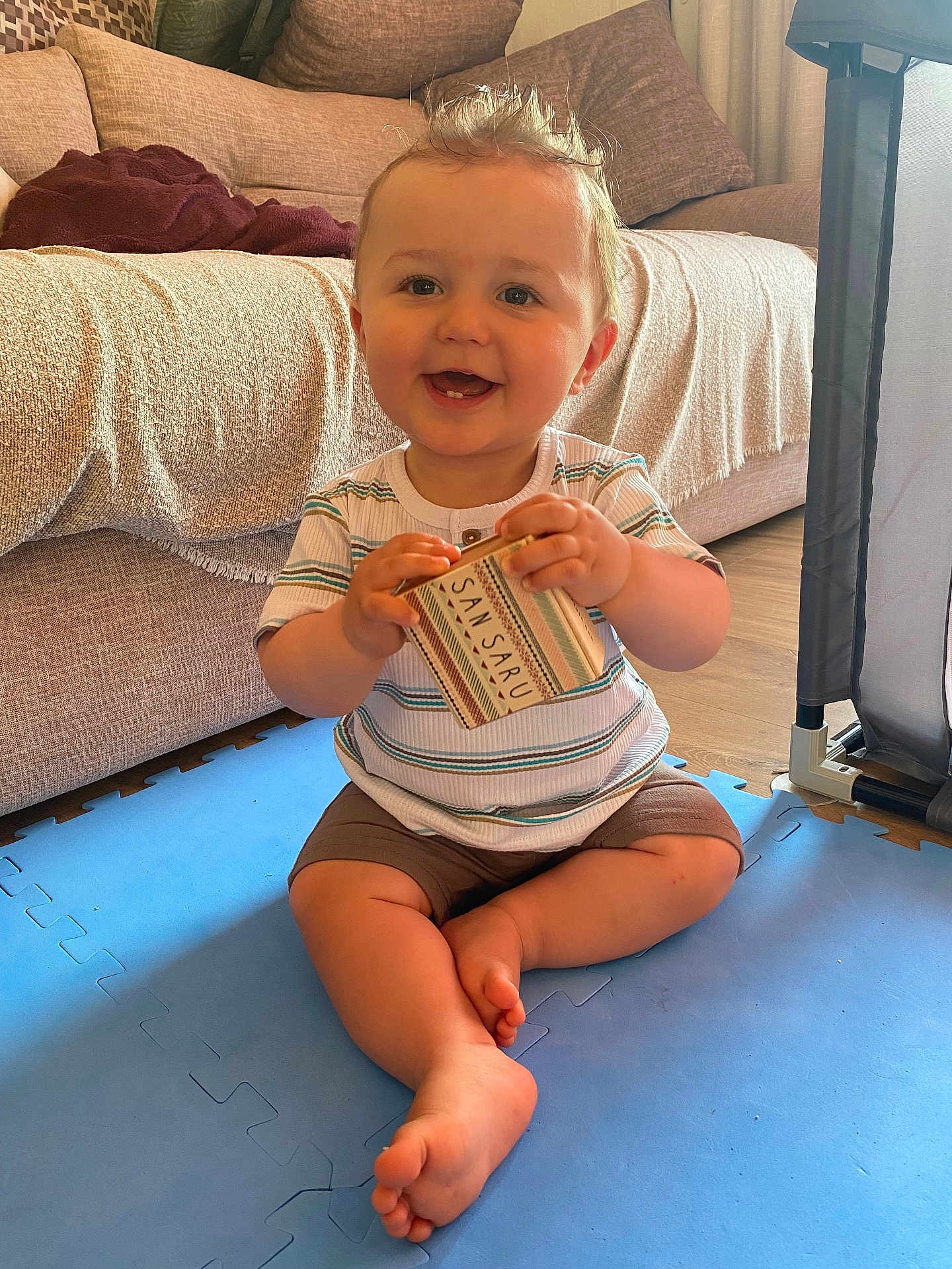 Andrea participe au concours pour gagner de l'argent avec cette photo : baby, child, smiling, indoor, play_mat, box, striped_shirt, shorts, barefoot, couch, pillow, floor, wooden_floor, happy, sitting, person, toddler, home, toy, cute