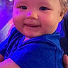 baby, child, infant, smiling, portrait, blue_shirt, curly_hair, face, eyes, cheeks, being_held, adult_hands, indoor, colorful_lighting, purple_lighting, blue_lighting, close_up, happy, photography, clothing