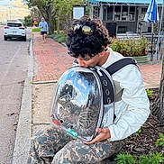 Zarco joined the competition — help win amazing prizes! cat, tabby_cat, pet_carrier, backpack, transparent, person, young_person, curly_hair, sunglasses, white_jacket, camouflage_pants, sneakers, outdoor, street, sidewalk, brick_pavement, cafe, bench, tree, cloudy_sky