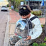 cat, tabby_cat, pet_carrier, backpack, transparent, person, young_person, curly_hair, sunglasses, white_jacket, camouflage_pants, sneakers, outdoor, street, sidewalk, brick_pavement, cafe, bench, tree, cloudy_sky