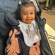 Giulia participe au concours pour gagner de l'argent avec cette photo : baby, smiling, child, stroller, bib, messy_face, hair_clip, seatbelt, wooden_floor, sunlight, outdoor, happy, infant, cute, person, clothing, hand, legs, black_stroller, daylight