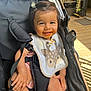 baby, smiling, child, stroller, bib, messy_face, hair_clip, seatbelt, wooden_floor, sunlight, outdoor, happy, infant, cute, person, clothing, hand, legs, black_stroller, daylight