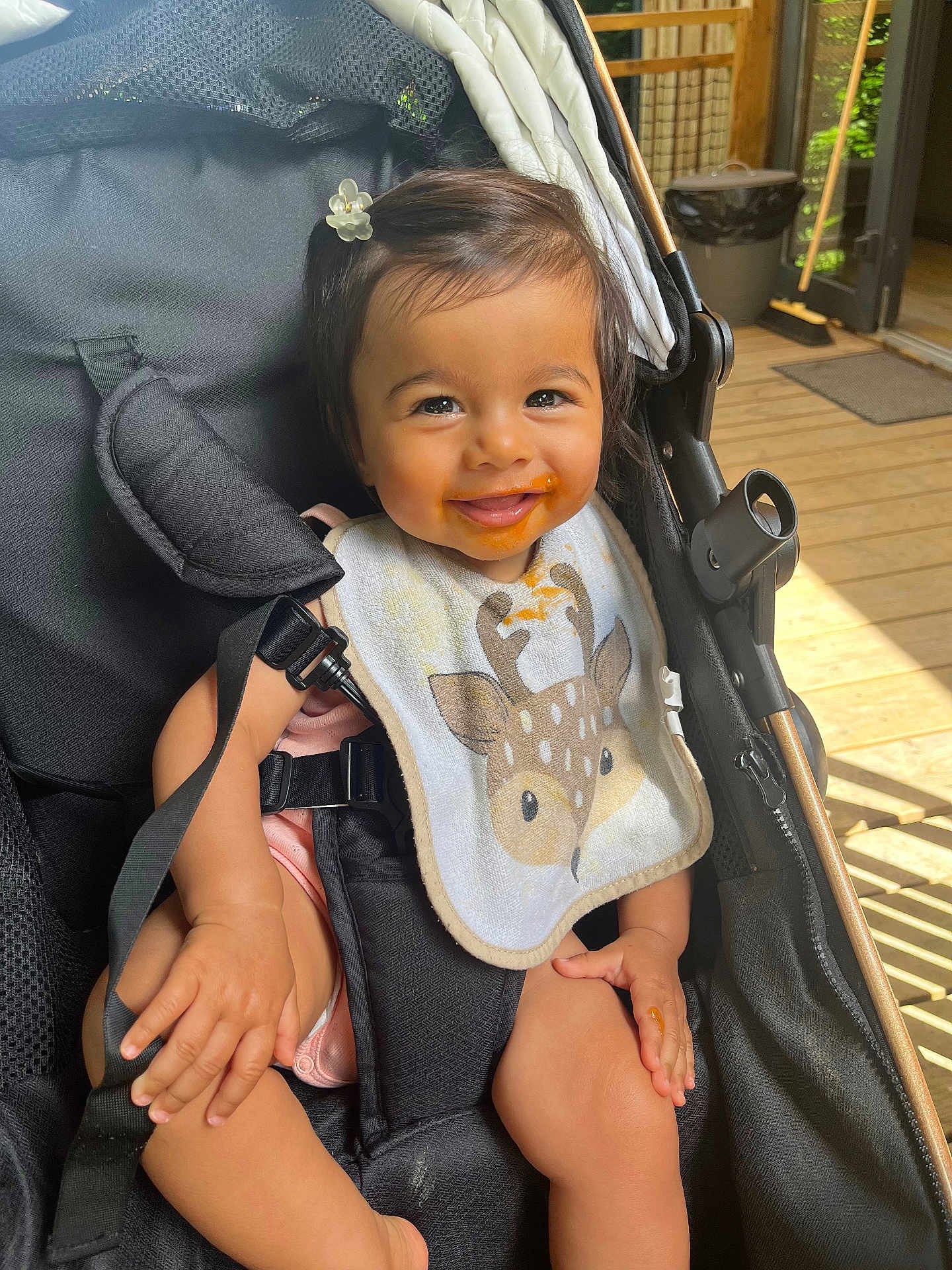 Giulia participe au concours pour gagner de l'argent avec cette photo : baby, smiling, child, stroller, bib, messy_face, hair_clip, seatbelt, wooden_floor, sunlight, outdoor, happy, infant, cute, person, clothing, hand, legs, black_stroller, daylight
