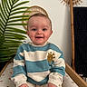 baby, child, smiling, sweater, striped_clothing, sitting, cushion, leaf, plant, indoor, decor, mirror, wood, pattern, happy, portrait, face, hand, cozy, cute