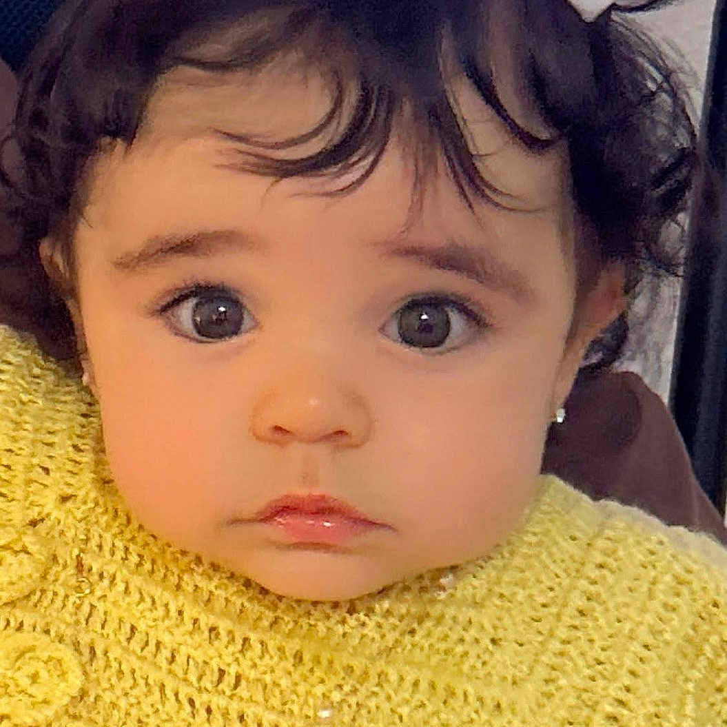 Aisha is registered to the contest to win money with this photo: adorable, baby, child, closeup, curly_hair, cute, earrings, expression, eyes, face, fashion, hair_bows, indoor, knitted_top, person, portrait, sitting, toddler, yellow_clothing, young_child