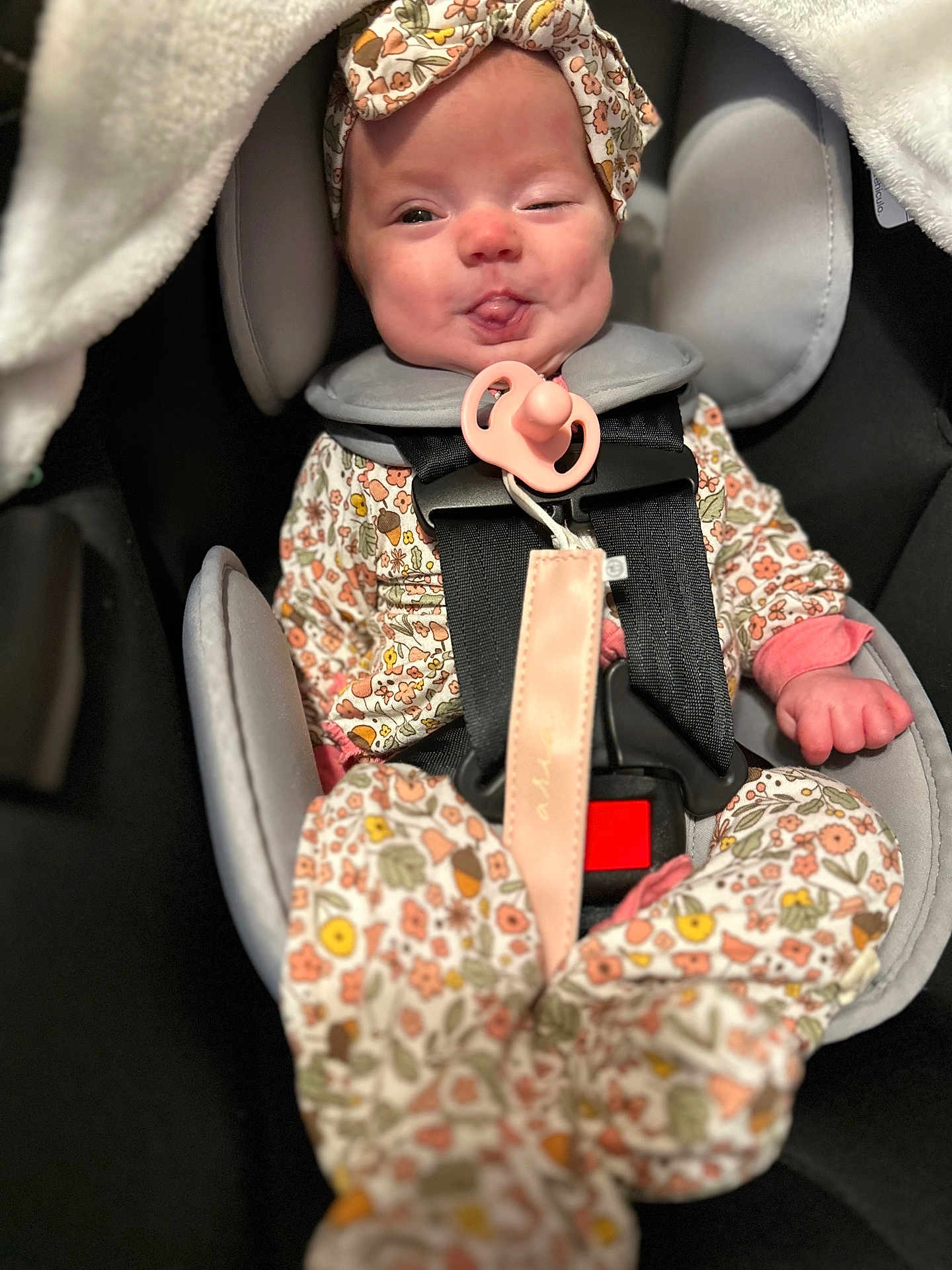 Aria is registered to the contest to win money with this photo: baby, infant, car_seat, pacifier, onesie, headband, floral_pattern, child, cute, tongue_out, smiling, seatbelt, soft_blanket, indoors, person, child_seat, baby_clothing, close_up, cute_expression, happy