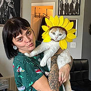Dublin is registered to the contest to win money with this photo: cat, chair, clothing, earrings, expression, framed_photo, furniture, ginger_cat, holding, indoor, living_room, person, pet, playful, portrait, ring, sunflower_costume, tattoos, wall_art, white_cat
