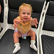 Amara-May is registered to the contest to win money with this photo: toddler, child, baby, smiling, reaching, chair, waiting_area, public_space, blond_hair, pacifier, socks, clothing, seated, cute, happy, indoor, person, young_child, expression, playful