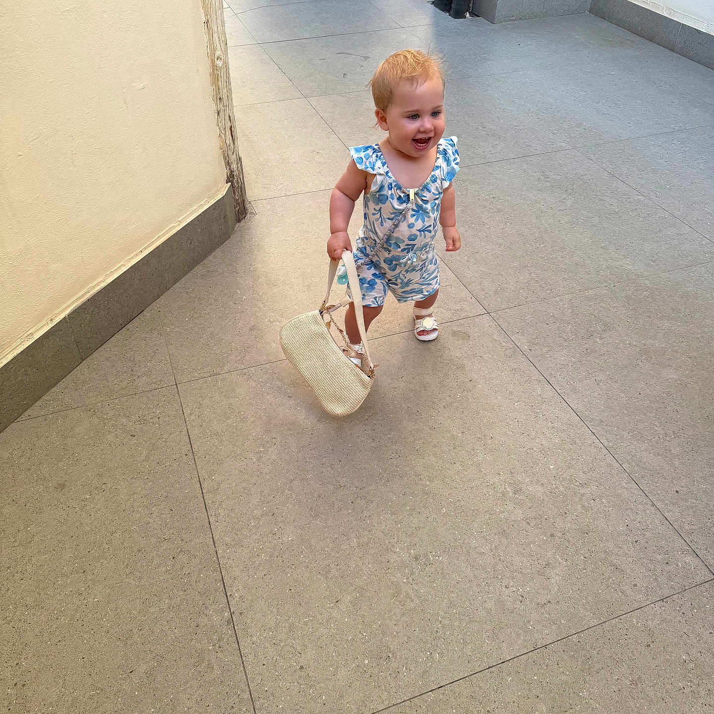 Amara-May is registered to the contest to win money with this photo: baby, child, corridor, cute, daylight, fashion, floor_tiles, footwear, hair, handbag, happy, outdoor, person, romper, sandals, small_bag, smiling, toddler, walking, wall