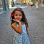 background_blur, child, cobblestone, curly_hair, daylight, dress, floral_pattern, flower, girl, happy, outdoor, people, portrait, posing, sandals, sidewalk, smile, street, summer, urban