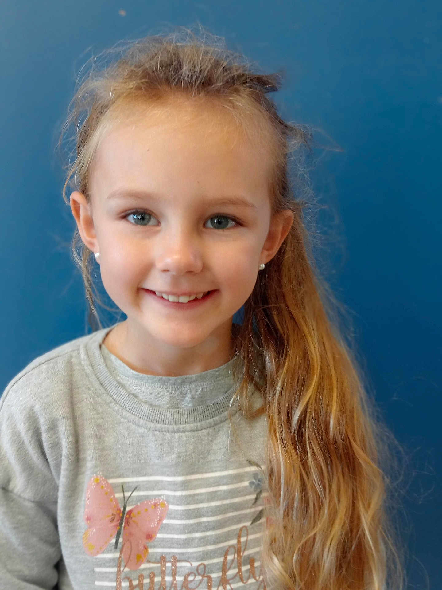 Cassie participe au concours pour gagner de l'argent avec cette photo : child, girl, smiling, long_hair, ponytail, grey_sweatshirt, butterfly, blue_background, earrings, casual_clothing, portrait, happy, face, young, cute, indoor, person, human, fashion, apparel