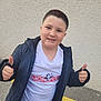 child, boy, thumbs_up, smile, white_tshirt, monopoly, jacket, short_hair, outdoor, wall, pavement, parking_line, casual_clothing, happy, posing, person, street, young, freckled_face, standing