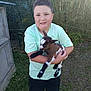 child, boy, goat, animal, pet, outdoor, grass, fence, green_shirt, black_pants, short_hair, smiling, holding, young, cute, sunlight, nature, farm, small_animal, happy
