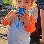 child, toddler, ball, playground, outdoor, sunlight, fence, swimwear, blonde_hair, curly_hair, playing, person, orange_sleeves, blue_shorts, toy, happy, daylight, summer, fun, smiling