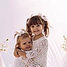 child, children, girl, toddler, hugging, smiling, white_dress, lace, sisters, family, portrait, happy, cute, flower, hair_bow, indoor, soft_light, twin_tails, tender, affection