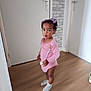 child, toddler, pink_clothing, pacifier, hair_bows, wooden_floor, door, wall, indoor, expression, standing, socks, person, home, hallway, curious, young_child, flooring, casual, portrait