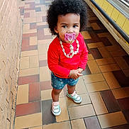 Kennedie participe au concours pour gagner de l'argent avec cette photo : toddler, child, indoor, pacifier, red_shirt, denim_shorts, mismatched_sneakers, afro_hair, shell_necklace, tile_floor, brick_wall, refrigerated_display, cute, standing, fashion, young_child, playful, casual_clothing, portrait, person