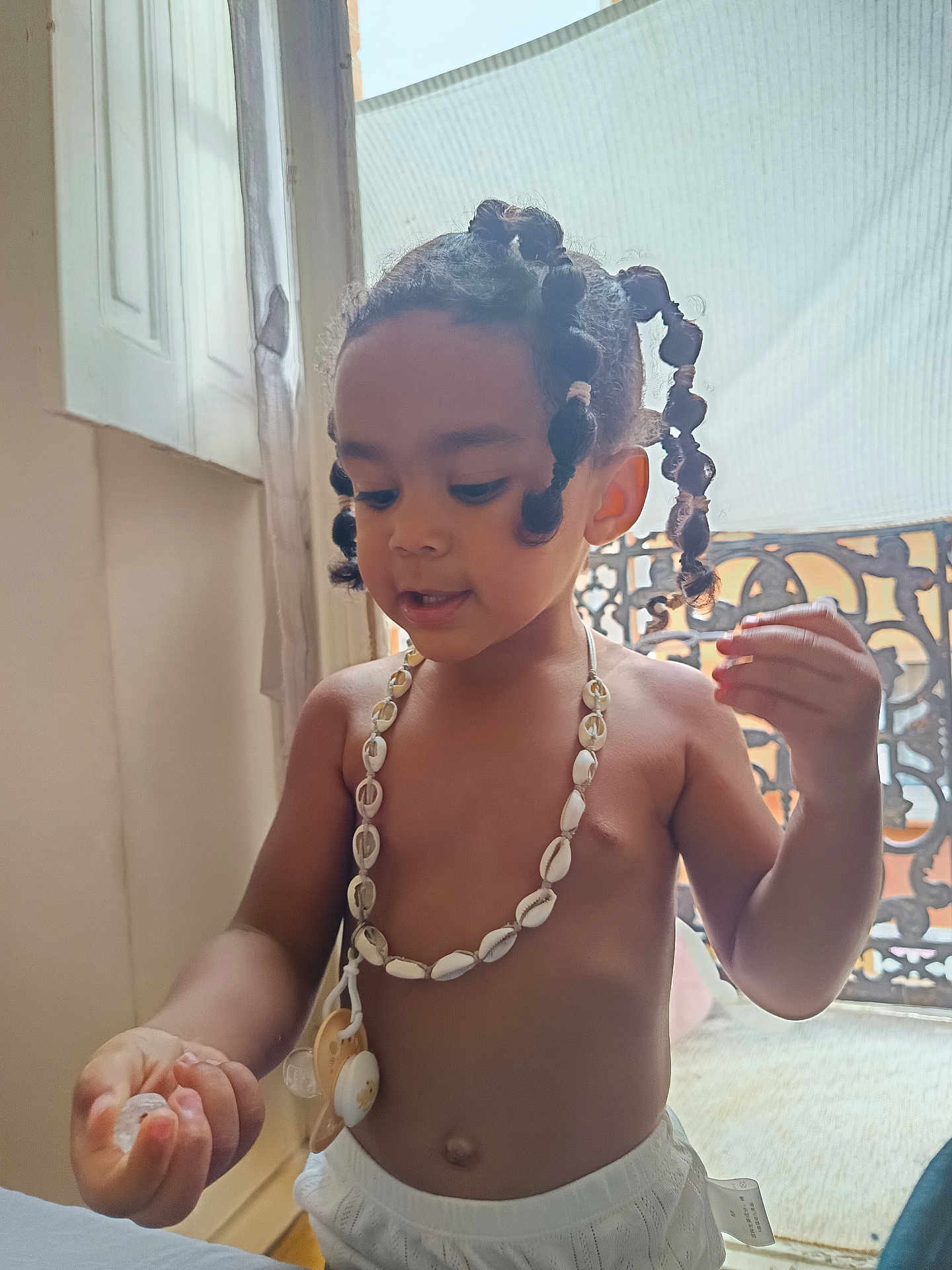 Kennedie participe au concours pour gagner de l'argent avec cette photo : child, toddler, indoor, hair, braids, shell_necklace, pacifier, shorts, window, light, curious, hands, face, skin, expression, natural_light, baby, person, young_child, decor