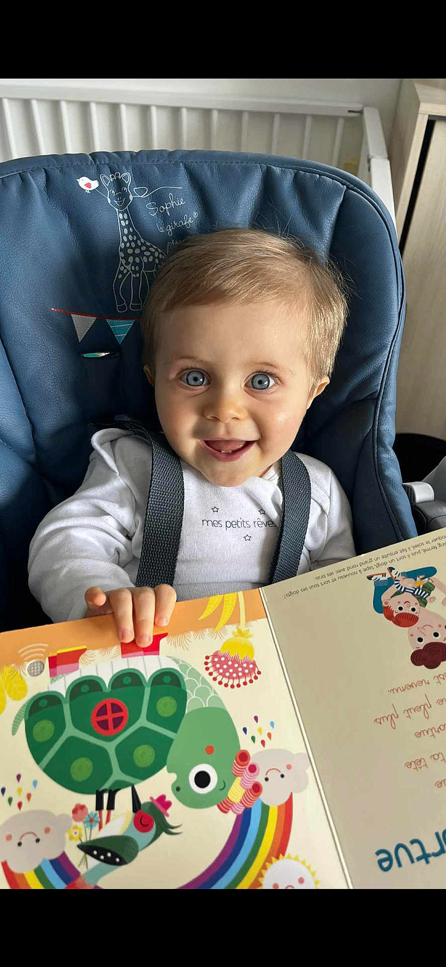 Oscar a rejoint le concours — aidez-le/la à gagner de superbes lots ! toddler, child, smiling, blue_high_chair, book, reading, illustration, turtle, rainbow, indoors, happy, cute, face, eyes, clothing, seatbelt, furniture, storytime, playful, colorful