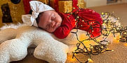 Amelie joined the competition — help win amazing prizes! baby, sleeping, red_dress, white_bow, christmas_lights, holiday, present, gift_box, decorations, carpet, pillow, star_shape, cozy, festive, indoors, newborn, cute, peaceful, soft_texture, holiday_season