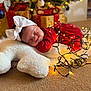 baby, sleeping, red_dress, white_bow, christmas_lights, holiday, present, gift_box, decorations, carpet, pillow, star_shape, cozy, festive, indoors, newborn, cute, peaceful, soft_texture, holiday_season