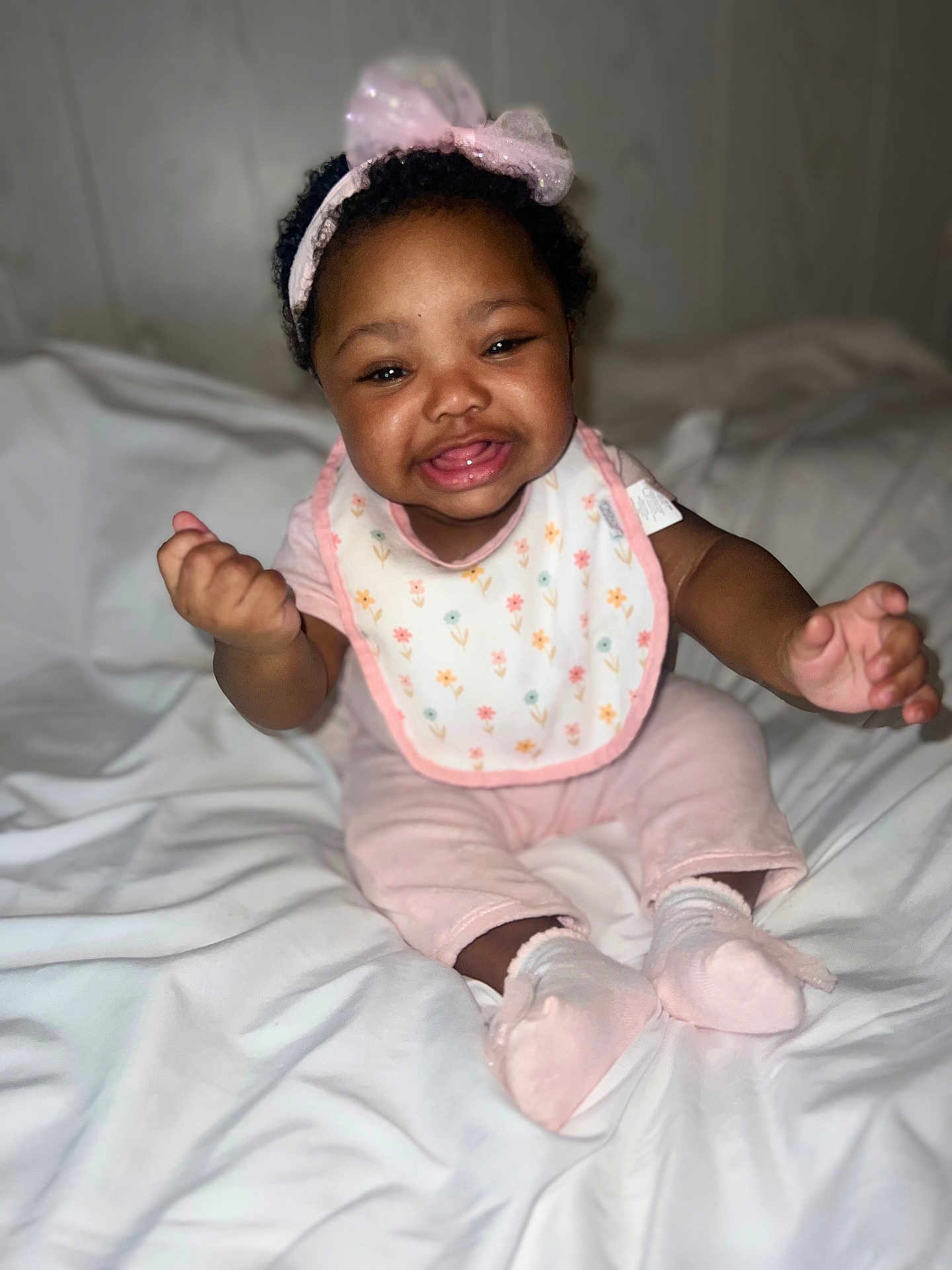 Ariyah is registered to the contest to win money with this photo: baby, child, smiling, pink_headband, bib, pink_clothing, socks, bed, white_bedspread, indoor, portrait, happy, cute, infant, person, young_child, sitting, reaching, face, curly_hair