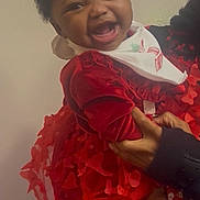 Ariyah is registered to the contest to win money with this photo: baby, child, smiling, red_dress, boots, bib, indoor, holding, person, happy, cute, textured_clothing, portrait, infant, hands, fashion, celebration, cheerful, face, closeup