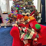 Meeyahanna Young is registered to the contest to win money with this photo: baby, child, plaid_dress, red_shoes, red_headband, plush_toy, reindeer_plush, elmo_plush, christmas_tree, holiday_decorations, festive, indoor, carpet, window, curtains, smiling, sitting, red_chair, holiday_season, happy