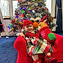 baby, child, plaid_dress, red_shoes, red_headband, plush_toy, reindeer_plush, elmo_plush, christmas_tree, holiday_decorations, festive, indoor, carpet, window, curtains, smiling, sitting, red_chair, holiday_season, happy