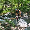 cap, creek, crouching, dog, forest, happy_dog, harness, leash, man, nature, outdoors, river, rock, rocks, shadows, smile, stream, sunlight, trees, water
