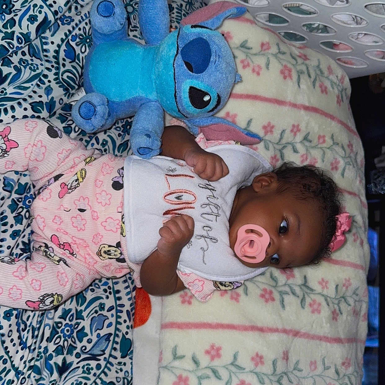 Makenzie joined the competition — help win amazing prizes! baby, bib, blanket, blue, child, comfort, cute, floral, indoors, infant, laundry_basket, lying_down, pacifier, pajamas, patterned_sheet, pink, plush_toy, sleepwear, stitch, toy