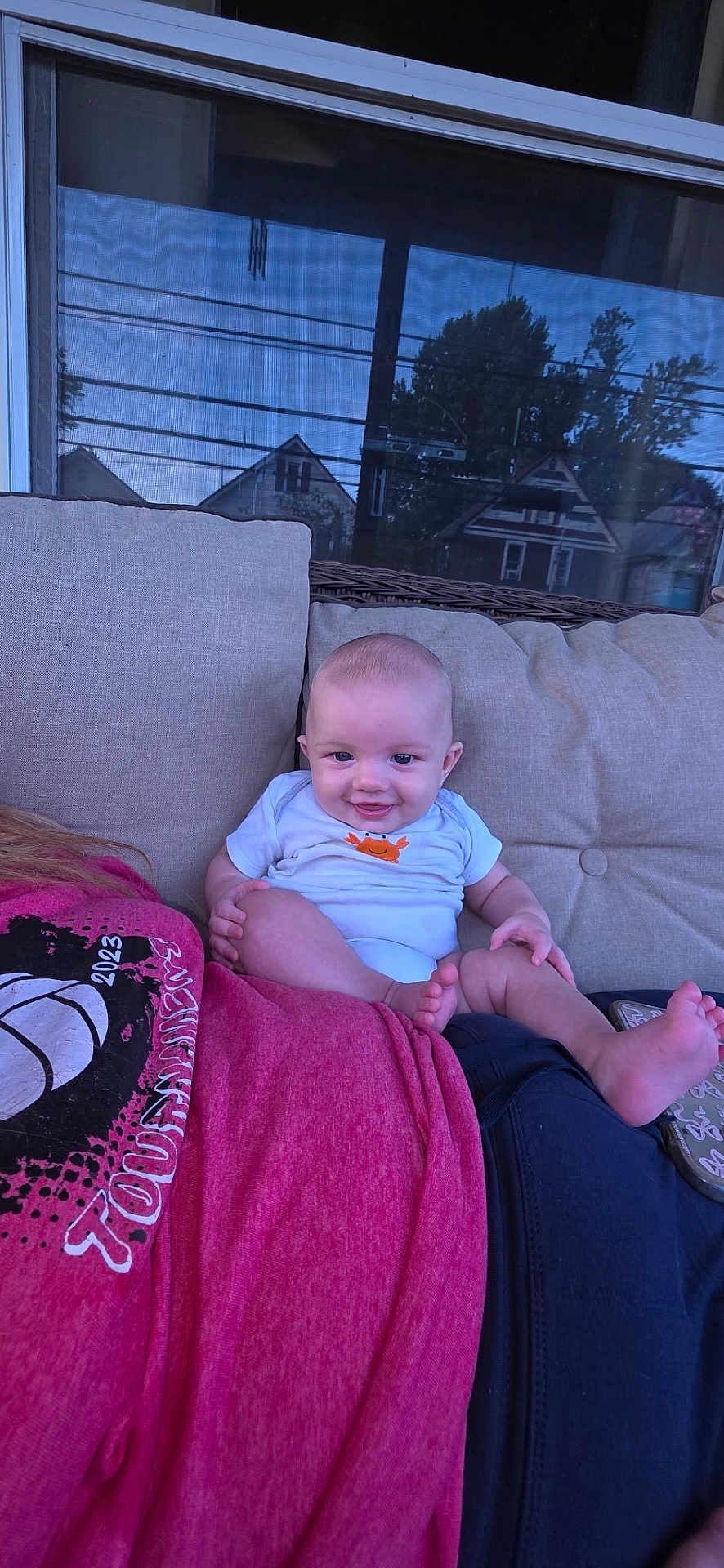 Luca is registered to the contest to win money with this photo: baby, smile, infant, onesie, person, couch, window, reflection, house, tree, outdoor, shirt, red_shirt, skin, foot, hand, relaxing, seated, home, casual